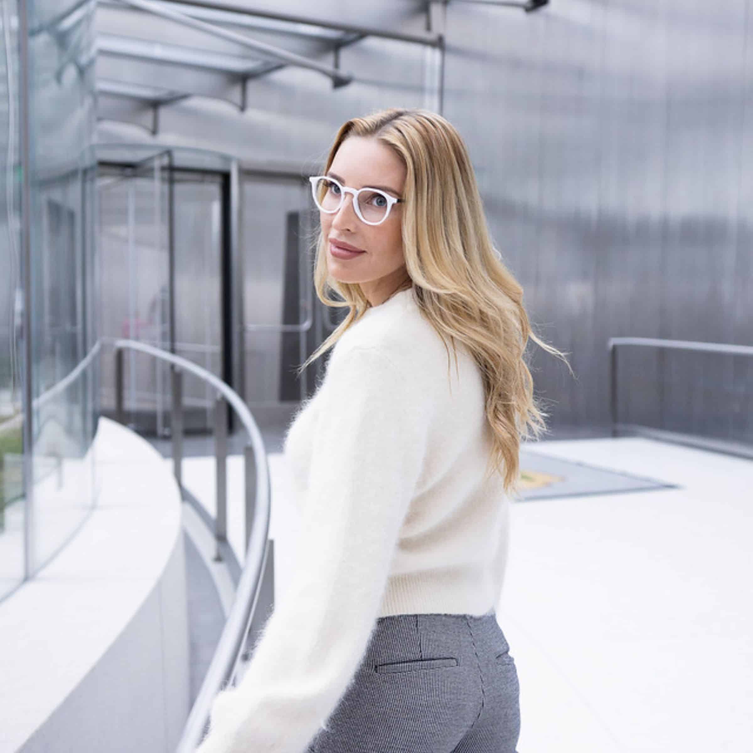 Woman in white sweater and gray pants wearing white-framed glasses, looking over her shoulder